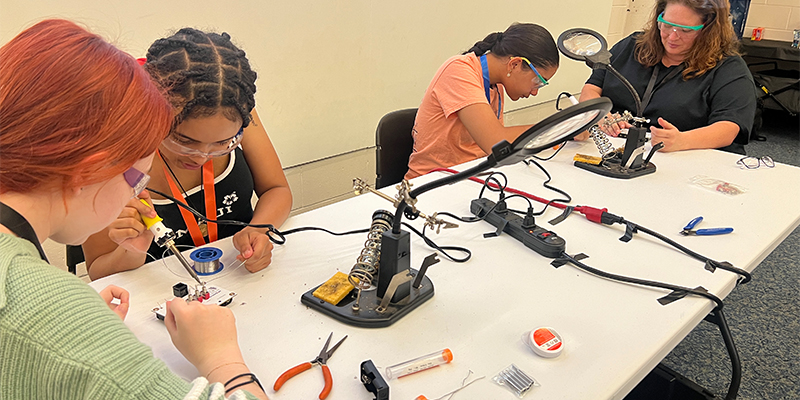 Three teenage girls building/soldering the ROV with instructor watching