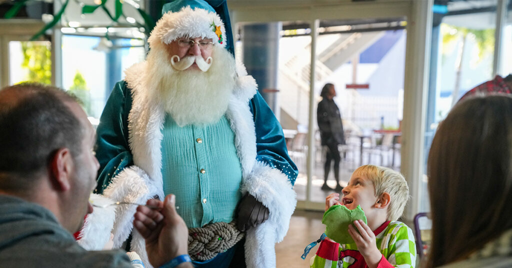 Little boy eating a pancake while smiling up at Santa wearing a blue suit while parents watch at The Florida Aquarium