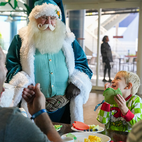 Little boy eating a pancake while smiling up at Santa wearing a blue suit while parents watch at The Florida Aquarium