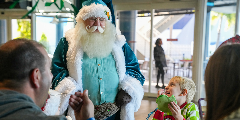 Little boy eating a pancake while smiling up at Santa wearing a blue suit while parents watch at The Florida Aquarium