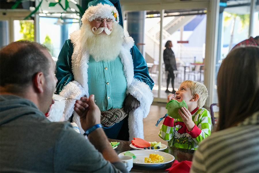 Little boy eating a pancake while smiling up at Santa wearing a blue suit while parents watch at The Florida Aquarium